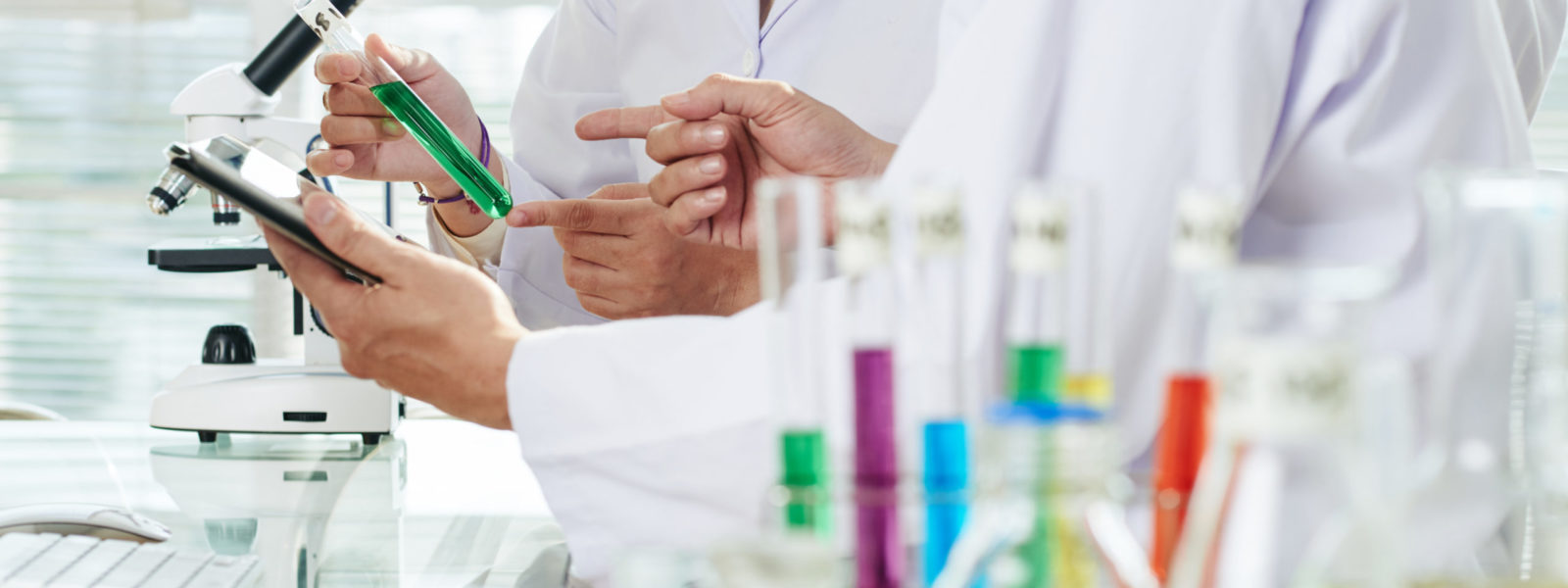 Close-up shot of unrecognizable chemists wearing white coats examining content of test tube and taking necessary notes with help of digital tablet while carrying out experiment